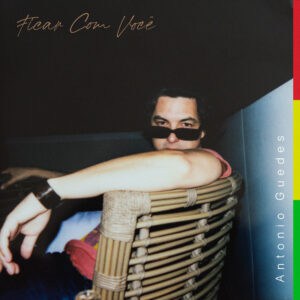 Stylish man wearing sunglasses, seated with arm resting on woven chair, in a modern indoor setting with dark background and minimalist decor.